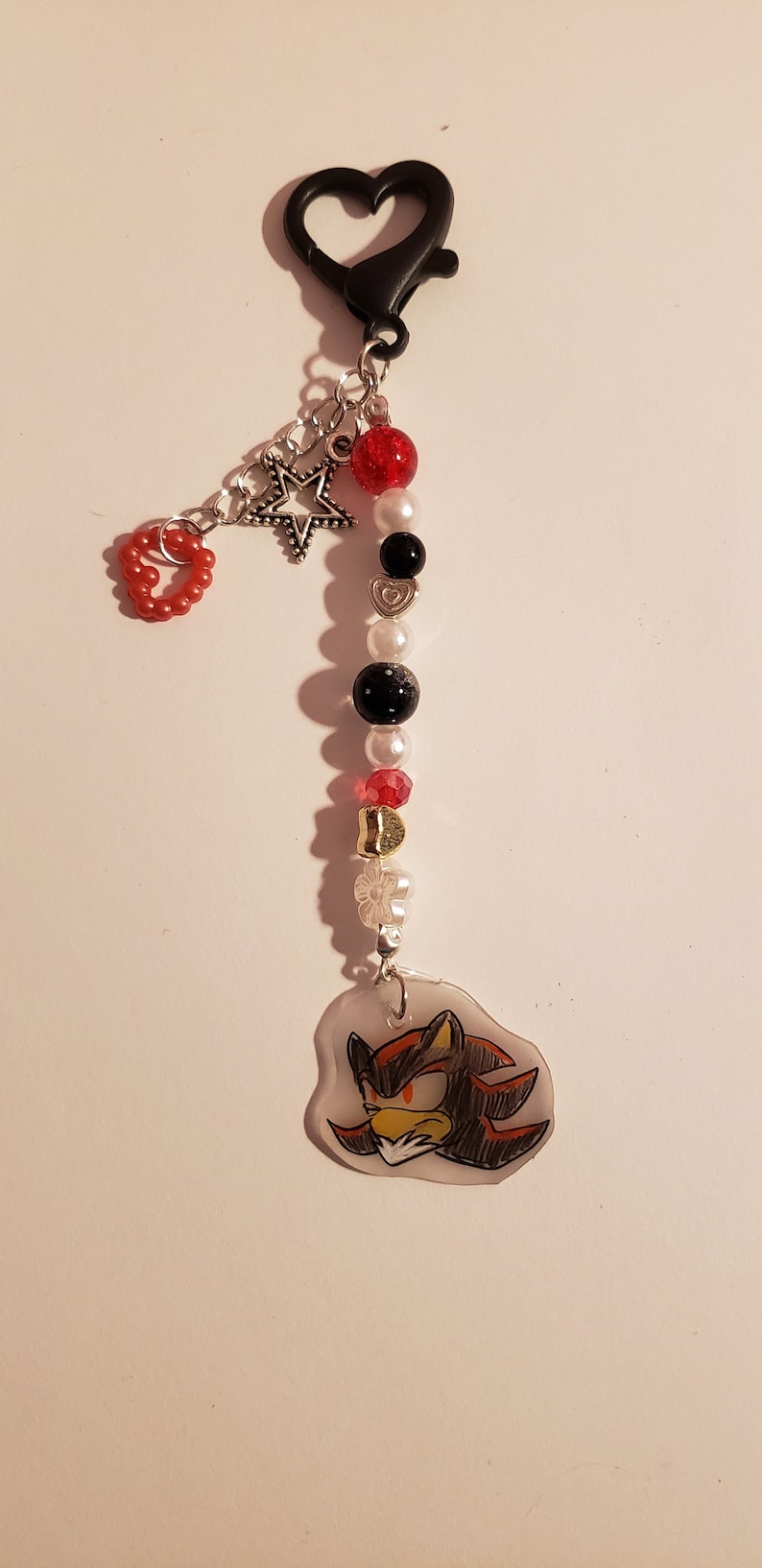 May include: A black keychain with a red heart, a silver star, and a clear charm with a drawing of Shadow the Hedgehog. The keychain is decorated with red, white, and black beads.