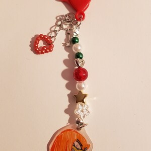 May include: A red heart-shaped keychain with a silver chain, a key charm, and a series of beads. The beads are green, white, and red with glitter. The keychain also features a charm with a cartoon image of a red-haired character with a yellow nose.