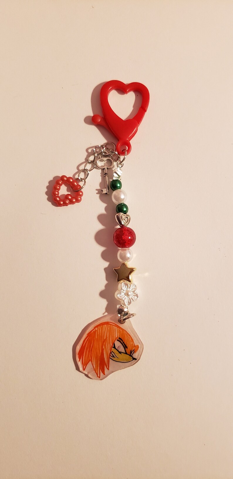 May include: A red heart-shaped keychain with a silver chain, a key charm, and a series of beads. The beads are green, white, and red with glitter. The keychain also features a charm with a cartoon image of a red-haired character with a yellow nose.