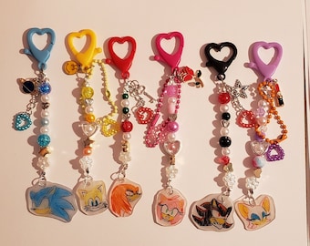 May include: Seven colorful keychains with heart-shaped clips and charms. Each keychain features a different character from the Sonic the Hedgehog franchise, including Sonic, Tails, Knuckles, Amy, Shadow, and Rouge. The charms include hearts, stars, and other decorative elements.