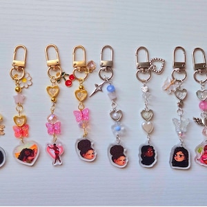 May include: A collection of keychains with gold-colored hardware. Each keychain features a charm with a portrait of a woman, along with decorative elements like butterflies, hearts, stars, and beads in various colors, including pink, gold, and clear.
