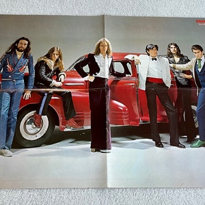 May include: A vintage red car with six men standing around it. The men are wearing various styles of clothing, including a blue jacket with a red and white pattern, a brown leather jacket, a black suit, a white shirt with a black bow tie, a blue shirt with a white vest, and a brown jacket with a blue vest. The car has a white steering wheel and whitewall tires.