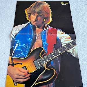 Puede incluir: Un póster vintage que muestra a un hombre tocando una guitarra eléctrica de cuerpo hueco. Lleva una chaqueta de satén azul con ribetes blancos y una correa de guitarra roja. El póster tiene el texto "SUPER POSTER" y "Andy Gibb" en la esquina superior derecha.