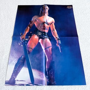 May include: A vintage poster featuring a muscular figure with long blonde hair, clad in black leather attire, holding a sword and a futuristic gun. The poster has a blue and purple background with the text "OKE.J" in the upper right corner.