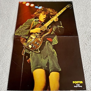 Op de afbeelding: Poster met Angus Young van AC/DC die gitaar speelt. De afbeelding toont Young in een groene fluwelen outfit, die een bruine elektrische gitaar bespeelt. De poster heeft een donkere achtergrond met podiumverlichting. Tekst op de poster luidt "POSTER AC/DC Angus Young."
