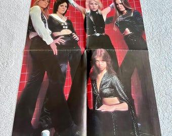 May include: A vintage poster featuring a group of women in 1970s attire. The women are posing in front of a red grid background. They are wearing black pants, tops, and jackets with silver accents. The poster is folded.