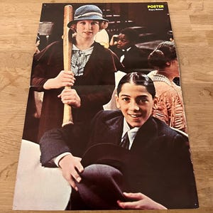 Può includere: Un poster vintage con una scena del film "Bugsy Malone". L'immagine mostra due bambini, uno con una mazza da baseball e l'altro con un cappello, con altre figure sullo sfondo. Il poster ha uno schema di colori marrone e rosso.