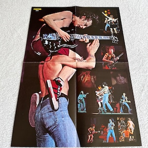 Op de afbeelding: Een gevouwen poster met meerdere afbeeldingen van een rockband die optreedt op het podium. De hoofdafbeelding toont een muzikant die een zwarte gitaar bespeelt. De poster heeft het woord "POSTER" bovenaan.