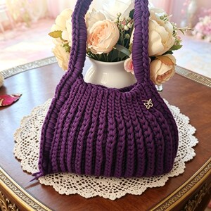 Può includere: Una borsa all'uncinetto viola scuro fatta a mano, con un manico intrecciato e un piccolo ciondolo a farfalla. La borsa è appoggiata su un centrino di pizzo bianco su un tavolo di legno. Un vaso di fiori è sullo sfondo.