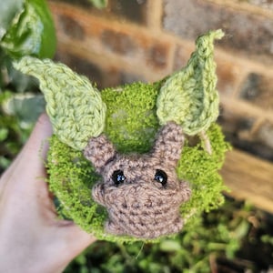 May include: A handcrafted crochet woodland creature with a brown face, ears, and eyes, and green leaves. The creature is set in a bed of green moss. The item is held in a person's hand.
