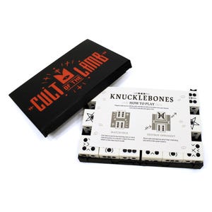 May include: A Cult of the Lamb Knucklebones board game set featuring a black box with red text, a white instruction card titled Knucklebones How to Play, and a collection of white dice with black symbols arranged in a black plastic tray.