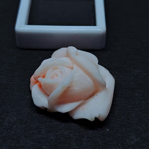 Op de afbeelding: Een gesneden lichtroze edelsteen in de vorm van een roos met delicate gelaagde bloemblaadjes en een gladde afwerking. De roos is geplaatst voor een klein, vierkant wit displaydoosje met een zwarte binnenkant.