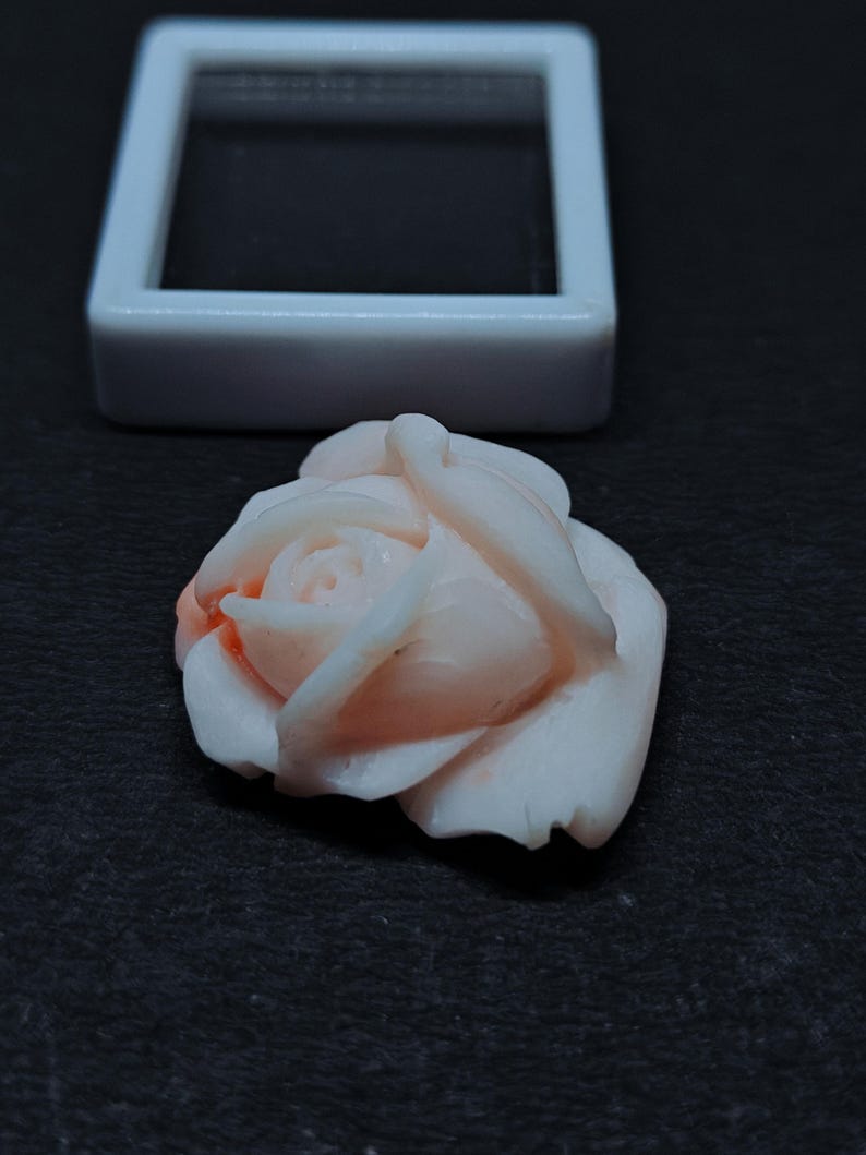 Op de afbeelding: Een gesneden lichtroze edelsteen in de vorm van een roos met delicate gelaagde bloemblaadjes en een gladde afwerking. De roos is geplaatst voor een klein, vierkant wit displaydoosje met een zwarte binnenkant.