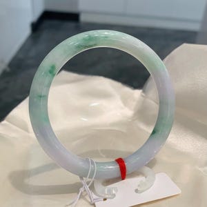 May include: A translucent jade bangle bracelet with a mix of pale green and lavender hues. The smooth, polished surface reflects light, showcasing the natural variations in color. A small red string and a white tag are attached to the bracelet.