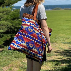 May include: A person wearing a grey shirt and black shorts is carrying a large tote bag with a colorful geometric pattern. The bag has a brown leather strap and a fringe detail. The person is standing in a grassy field with a body of water in the background.