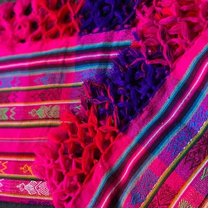 Puede incluir: Una manta tejida de color rosa y morado con rayas coloridas y patrones intrincados. La manta tiene flecos en todos los lados.