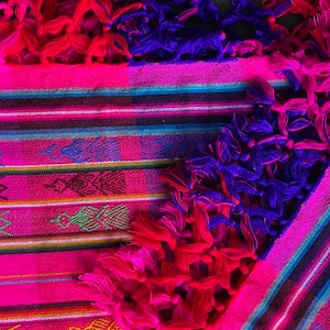 May include: A pink and blue woven textile with fringe. The textile has a striped pattern with geometric designs.