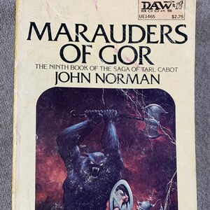 May include: Vintage paperback book titled "Marauders of Gor" by John Norman. The cover art depicts a fierce, dark blue monster wielding an axe, battling a warrior in purple armor. The book is from DAW Books.