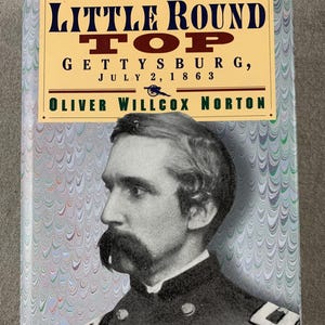 May include: A hardcover book titled "The Attack & Defense of Little Round Top Gettysburg, July 2, 1863" by Oliver Willcox Norton. The book cover features a portrait of a man with a mustache and a decorative marbled background.