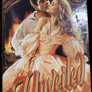 May include: Book cover for "Unveiled" by Jenni Licata. The cover features a couple embracing in front of a fireplace. The woman wears a peach-colored gown, and the title is in a stylized font.