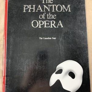 May include: A black programme cover with the title "The Phantom of the Opera" in white lettering. "The Canadian Tour" is printed below the title. A white mask is on the lower right corner. The spine is red.