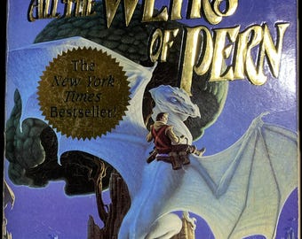 May include: Book cover for "All the Weyrs of Pern" by Anne McCaffrey. The cover features a white dragon with a rider soaring above a mountainous landscape. The title and author's name are in gold lettering against a blue background. A gold seal reads "The New York Times Bestseller!"