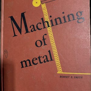 May include: A vintage book titled "Machining of metal" by Robert E. Smith. The cover is a reddish-brown color with black text and a yellow illustration of a spring and circle. The book appears to be an older edition.
