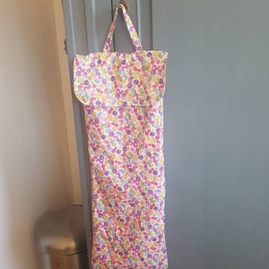 Puede incluir: Una bolsa de tela blanca con un patrón floral en tonos de rosa, amarillo, morado y verde. La bolsa tiene un asa larga y está colgada de una puerta.