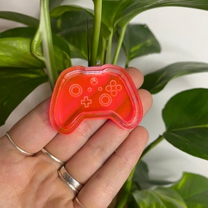 May include: A bright pink plastic video game controller keychain. The controller has a raised design of the buttons and a clear plastic background.