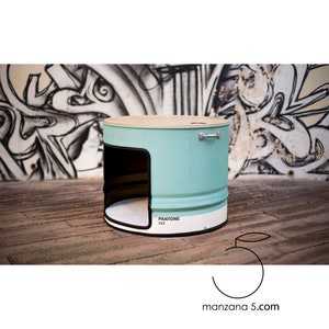 Puede incluir: Una cama cil&iacute;ndrica para mascotas en color turquesa claro, con tapa de madera y base blanca. La cama tiene una abertura rectangular y un asa. El c&oacute;digo de color Pantone 332 es visible. Dise&ntilde;ada para mascotas.