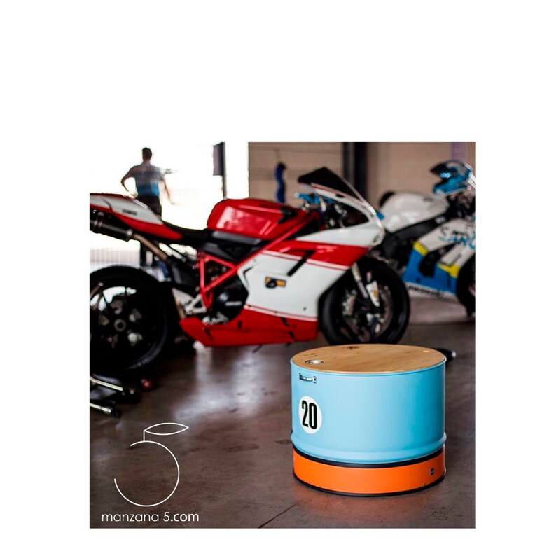 Puede incluir: Una mesa auxiliar en forma de barril azul claro y naranja con tapa de madera. El n&uacute;mero "20" se muestra en el lateral. Dos motocicletas est&aacute;n en el fondo. El sitio web "manzana 5.com" se muestra en la esquina inferior izquierda.