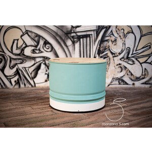 Puede incluir: Mesa redonda hecha de un barril reciclado. El cuerpo est&aacute; pintado de azul claro, con una base blanca y una tapa de madera natural. La mesa est&aacute; sobre un suelo de madera. El fondo presenta grafitis. Di&aacute;metro aproximado de 50 cm.
