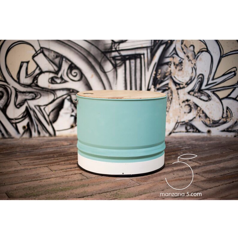 Puede incluir: Mesa redonda hecha de un barril reciclado. El cuerpo est&aacute; pintado de azul claro, con una base blanca y una tapa de madera natural. La mesa est&aacute; sobre un suelo de madera. El fondo presenta grafitis. Di&aacute;metro aproximado de 50 cm.