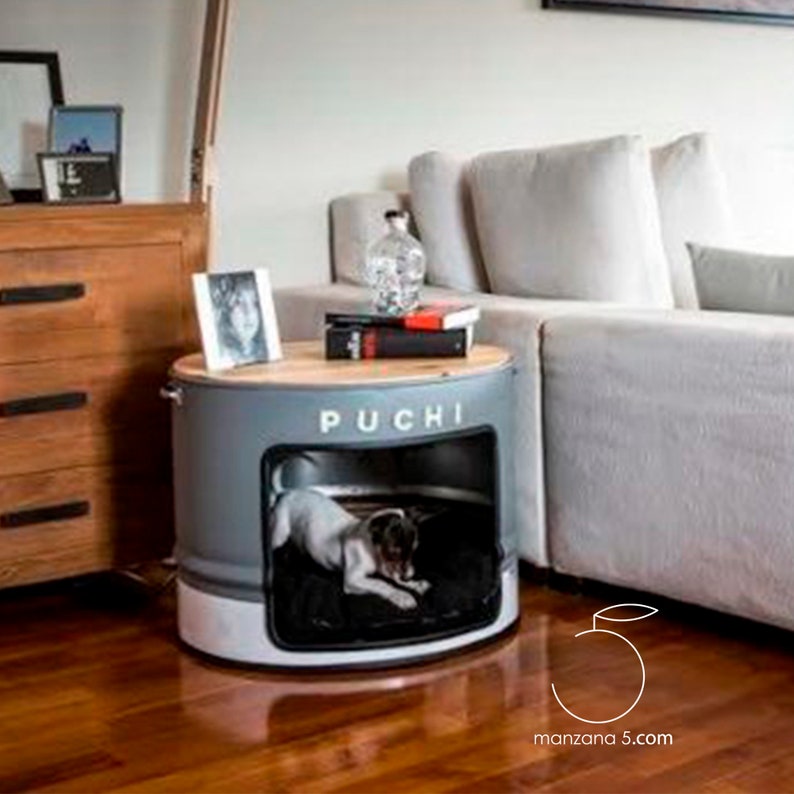 Puede incluir: Una cama para perros gris y blanca con el nombre "PUCHI" en letras blancas. La cama est&aacute; hecha de un tambor de metal reutilizado con una tapa de madera. Un perro peque&ntilde;o est&aacute; dentro de la cama sobre un coj&iacute;n negro. La cama est&aacute; al lado de un sof&aacute; gris claro.