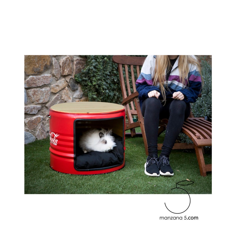 Puede incluir: Una cama roja para mascotas hecha de un barril de Coca-Cola reutilizado. La cama tiene una tapa de madera y un coj&iacute;n negro en el interior. Una mascota blanca y esponjosa est&aacute; dentro de la cama. El logotipo de Coca-Cola es visible en el lateral.