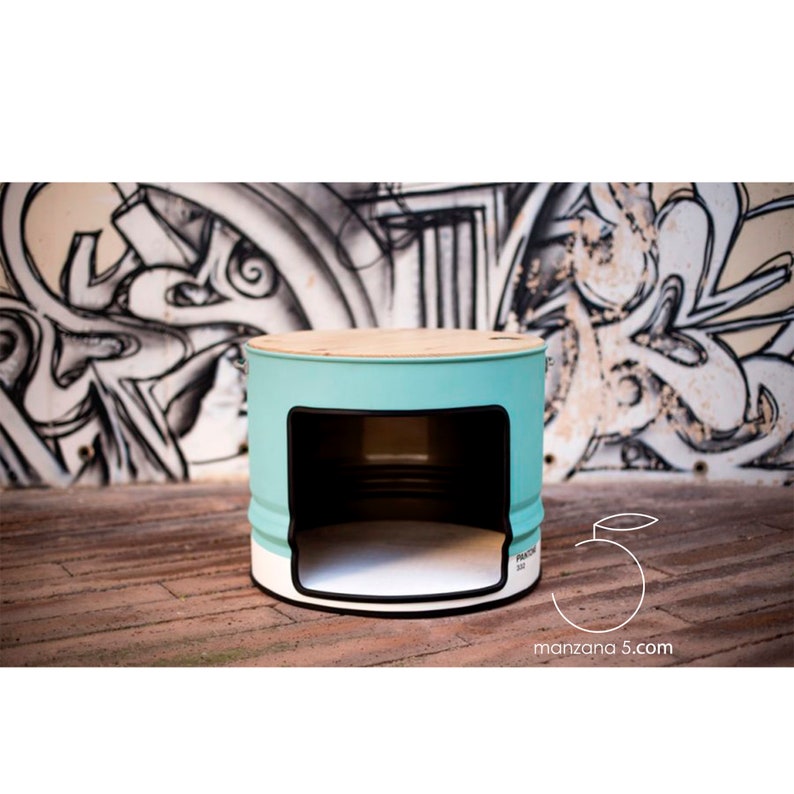 Puede incluir: Una cama para mascotas turquesa y blanca hecha de un tambor de metal reutilizado. La cama tiene una tapa de madera y una abertura rectangular. La cama est&aacute; pintada con el color Pantone 552. El fondo presenta graffiti.
