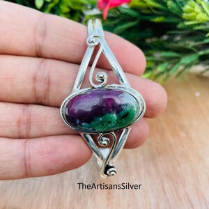 Puede incluir: Colgante de plata con una piedra ovalada en tonos morados y verdes. El colgante tiene un dise&ntilde;o calado con detalles en espiral. El texto "TheArtisansSilver" es visible en la parte inferior del colgante.