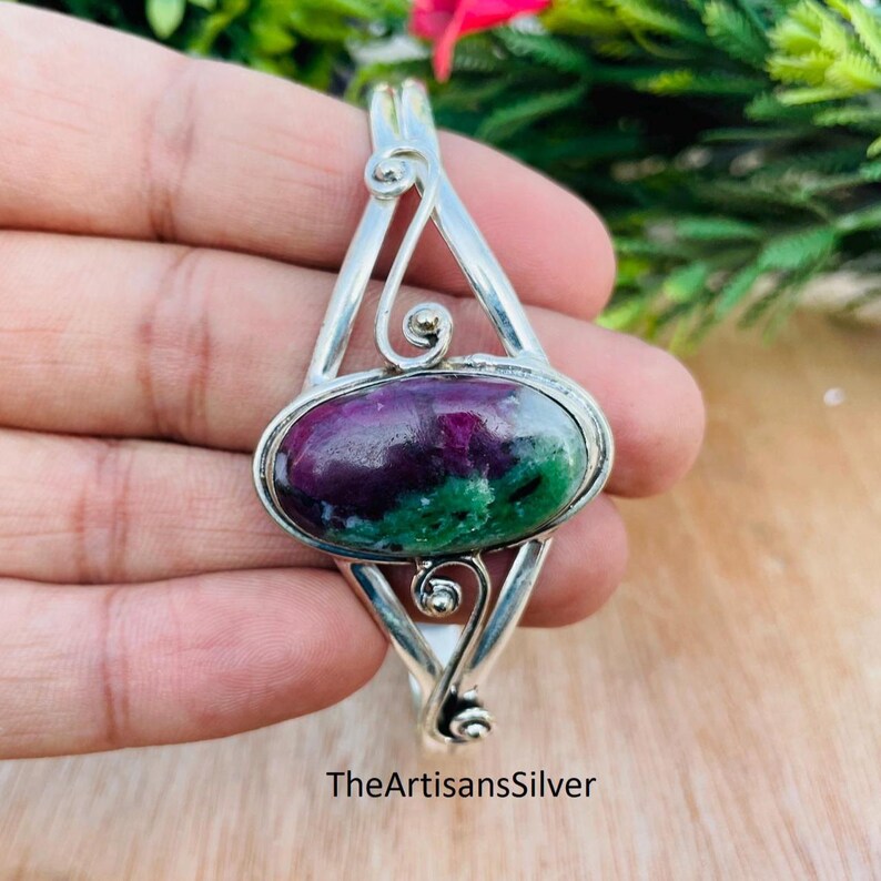 Puede incluir: Colgante de plata con una piedra ovalada en tonos morados y verdes. El colgante tiene un dise&ntilde;o calado con detalles en espiral. El texto "TheArtisansSilver" es visible en la parte inferior del colgante.