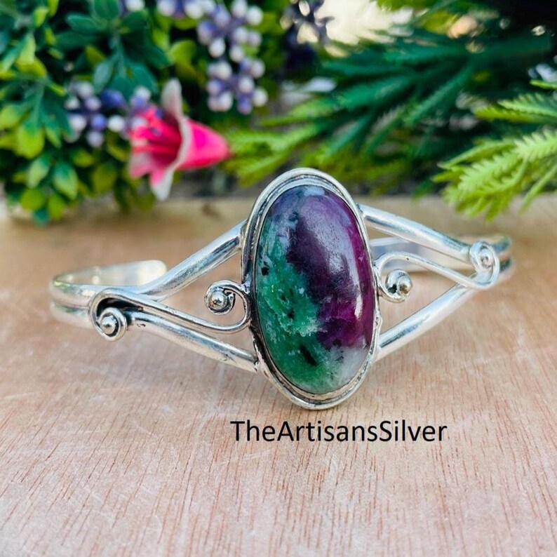 Puede incluir: Una pulsera de plata con una piedra preciosa ovalada. La piedra preciosa presenta una mezcla de colores rojo oscuro y verde. La pulsera tiene un dise&ntilde;o decorativo con remolinos y el texto "TheArtisansSilver".