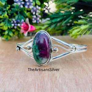 Puede incluir: Un brazalete de plata con una piedra preciosa ovalada en tonos rojo oscuro, verde y blanco. El brazalete tiene un dise&ntilde;o abierto con remolinos decorativos y el texto "TheArtisansSilver".