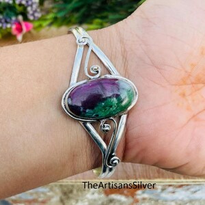 Puede incluir: Una pulsera de plata con una gema ovalada en tonos morados y verdes. La pulsera tiene un dise&ntilde;o abierto con remolinos decorativos. El texto "TheArtisansSilver" es visible en la parte inferior.