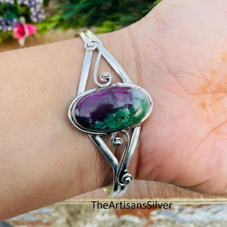 Puede incluir: Una pulsera de plata con una gema ovalada en tonos morados y verdes. La pulsera tiene un dise&ntilde;o abierto con remolinos decorativos. El texto "TheArtisansSilver" es visible en la parte inferior.