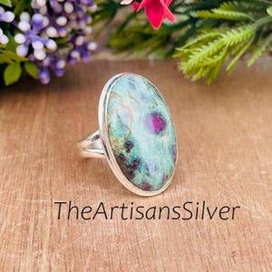 Pode incluir: Anel oval de prata com uma grande pedra preciosa azul-esverdeada clara com inclusões vermelhas e verde escuro. O anel é exibido em uma superfície de madeira. O texto "TheArtisansSilver" é visível.