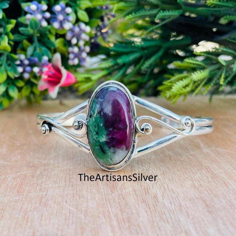 Puede incluir: Una pulsera de plata con una piedra preciosa ovalada. La piedra preciosa presenta tonos rojos oscuros y verdes. La pulsera tiene un dise&ntilde;o abierto con remolinos decorativos y el texto "TheArtisansSilver".