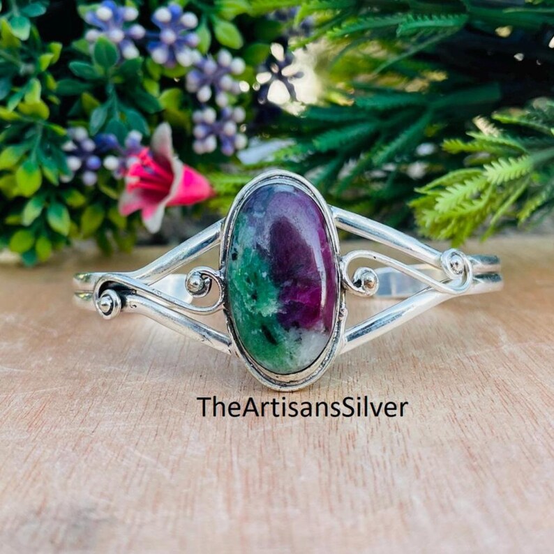 Puede incluir: Un brazalete de plata con una piedra preciosa ovalada en tonos rojo oscuro y verde. El brazalete tiene un dise&ntilde;o decorativo. El texto "TheArtisansSilver" es visible en la superficie.