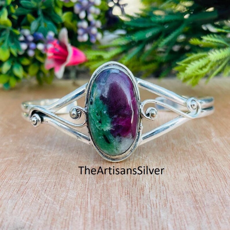 Puede incluir: Un brazalete de plata con una piedra preciosa ovalada en tonos rojos, verdes y blancos. El brazalete tiene un dise&ntilde;o decorativo y el texto "TheArtisansSilver" es visible en la parte inferior. Se encuentra sobre una superficie de madera.