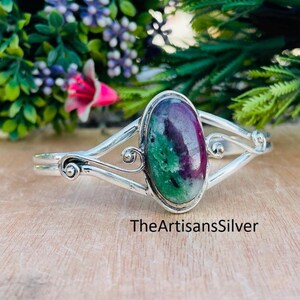 Puede incluir: Un brazalete de plata con una piedra preciosa ovalada en tonos verdes y granates. El brazalete tiene un dise&ntilde;o decorativo con espirales y curvas. El texto "TheArtisansSilver" es visible. Joyer&iacute;a artesanal.