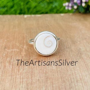 May include: A silver ring with a round, white centerpiece featuring a spiral design. The ring is set against a wooden surface, with the text "TheArtisansSilver" visible below the ring.