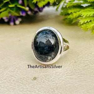 Puede incluir: Un anillo de plata con una piedra preciosa grande, ovalada y de color gris oscuro. La piedra preciosa tiene una superficie reflectante. El anillo está engastado en una banda de plata pulida. El texto "TheArtisansSilver" es visible en la parte inferior de la imagen.