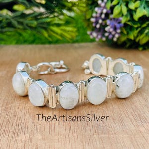 Puede incluir: Un brazalete de plata con piedras preciosas ovaladas de color blanco lechoso. El brazalete tiene un cierre seguro y se exhibe sobre una superficie de madera. El texto "TheArtisansSilver" es visible.